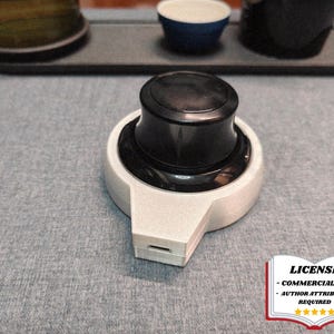 May include: A black and white computer mouse with a cylindrical top and a rectangular base. The mouse is on a gray surface. The image also includes a tray with cups and a license badge.