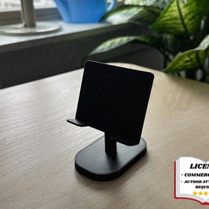 Può includere: Un supporto nero per smartphone con un supporto rettangolare e un piccolo bordo per tenere il telefono. Il supporto ha una base rettangolare arrotondata e un supporto verticale. Il supporto è su una superficie di legno chiaro.
