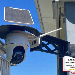May include: A white security camera with a black solar panel and a black mounting bracket. The camera has a dome shape and a white antenna. The bracket is attached to a pole. The sky is blue.