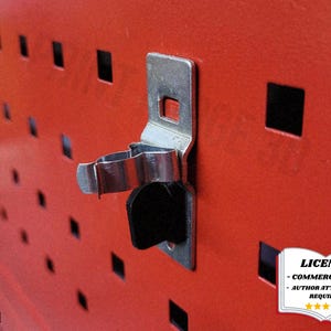 May include: Close-up of a red metal panel with square perforations and a silver-colored clip. The clip is attached to the panel with a black rubber piece. The panel has a license badge in the bottom right corner.