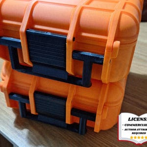 Puede incluir: Dos maletas rígidas apiladas de color naranja con cierres y bisagras negros. Las maletas están hechas de un material plástico duradero. La imagen incluye una insignia de licencia en la esquina inferior derecha.