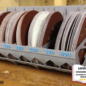 May include: A gray, multi-compartment organizer filled with various sizes of sanding discs. The discs are brown and tan, with blue numbers indicating grit sizes. The organizer is on a wooden surface. The text "LICENSE - COMMERCIAL USE - AUTHOR ATTRIBUTION REQUIRED" is visible.
