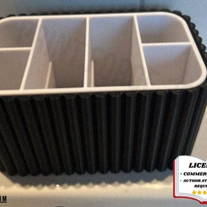 Puede incluir: Un organizador en blanco y negro con múltiples compartimentos. El exterior es negro con un diseño acanalado, y el interior es blanco. El organizador está diseñado para guardar varios artículos y es adecuado para uso doméstico o de oficina.