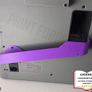 May include: A purple 3D-printed phone holder mounted on a grey computer. The holder is designed to hold a phone, with the text "PRINT FORGE" faintly visible on the computer. The image also includes the text "@CIMERA 3D".
