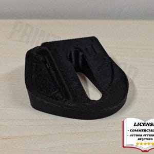 以下が含まれることがあります： 黒色の3Dプリントされたオブジェクトで、湾曲したデザインと中央の開口部があります。オブジェクトはテクスチャ加工された表面を持ち、明るい木製の背景に配置されています。ライセンス通知が右下に表示されています。