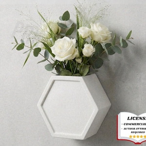 May include: A white, hexagonal wall vase filled with white roses, eucalyptus, and ornamental grasses. The vase is mounted on a light grey wall. The image includes a license badge in the bottom right corner.