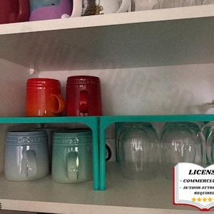 May include: A teal shelf organiser inside a white cupboard, holding various mugs in shades of red, blue, and green. Clear drinking glasses are also visible. The image includes a license notice.