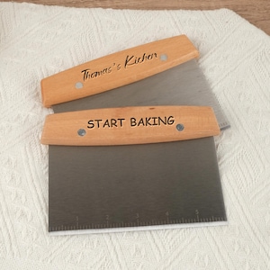 Op de afbeelding: Twee roestvrijstalen deegschrapers met houten handvatten. Op één handvat staat "Thomas's Kitchen", op de andere "START BAKING". De messen hebben meetmarkeringen. Op een witte, getextureerde ondergrond.