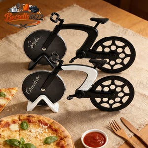 Peut inclure: Deux coupe-pizzas en forme de vélo. L'un est noir avec une base blanche et le nom "Charlotte" sur la roue. L'autre est noir avec une base noire et le nom "Sophia" sur la roue. Pizza et sauce au premier plan.