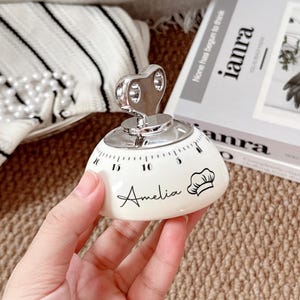 May include: A white dome-shaped kitchen timer with a metallic silver wind-up key on top. The timer features black numerical markings from 0 to 20, the name Amelia in cursive script, and a small black chef hat icon printed on the side surface.