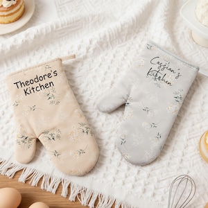May include: Two oven mitts with floral patterns and the text "Theodore's Kitchen" and "Cassian's Kitchen". One mitt is beige, the other is gray. Both have white and green floral designs.