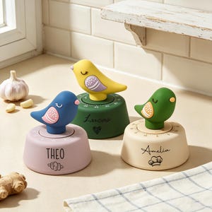 May include: Three round kitchen timers topped with bird figurines. The left timer is light pink with the name THEO and a croissant icon. The center timer is dark green with the name Lucas and a heart. The right timer is cream with the name Amelia and a chef hat icon.