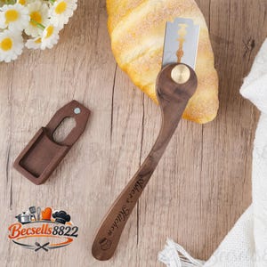 May include: A wooden bread lame with a metal blade and a protective sheath. The handle is engraved with "Asher's Kitchen". A croissant rests nearby on a wooden surface. The logo "Becsells8822" is in the lower left corner.