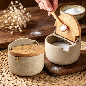 Op de afbeelding: Twee gespikkelde beige keramische zoutpotjes met draaibare bamboe deksels. Eén deksel is voorzien van een bloemenkransmotief met de naam Oliver erin gegraveerd. Een klein houten lepeltje schept wit zout in de open, met zout gevulde houder.