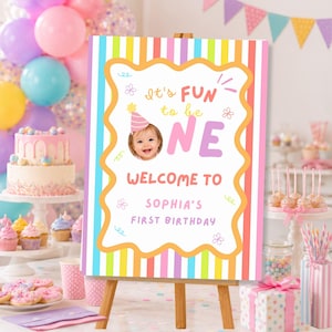 May include: A rectangular birthday welcome sign with a vertical striped border in pastel colors. The center features a white scalloped frame with the text It's FUN to be ONE, a photo of a baby in a party hat, and the text WELCOME TO SOPHIA'S FIRST BIRTHDAY.