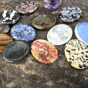 May include: A collection of polished, oval-shaped gemstones in various colors and patterns. The stones include a tiger's eye, a blue sodalite, a snowflake obsidian, and a red and white patterned stone. The gemstones are arranged on a dark, textured surface.