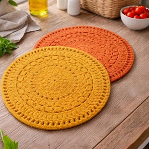 Puede incluir: Dos manteles individuales redondos de ganchillo en tonos amarillo y naranja descansan sobre una superficie de madera. Los manteles individuales tienen un aspecto texturizado con un borde decorativo. Otros elementos en el fondo incluyen una botella de aceite, saleros y pimenteros y una cesta de pan.