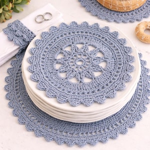 May include: A stack of white plates topped with a light blue crocheted placemat. The placemat has a circular design with a scalloped edge. A matching napkin ring and a napkin are visible on the left.