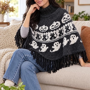 May include: A dark charcoal grey knit poncho featuring a high turtleneck collar and long fringe along the bottom hem. The garment displays a repeating white jacquard pattern of jack-o'-lanterns, black cats, and ghosts across the front surface.
