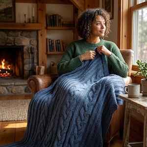 Puede incluir: Una persona sentada en un sillón de cuero, cubierta con una manta azul de punto trenzado. La persona lleva un suéter verde. Una chimenea con fuego ardiendo está en el fondo, creando un ambiente cálido.