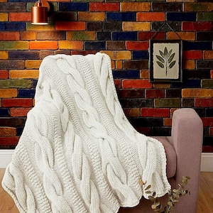 Puede incluir: Una manta de punto trenzado color crema sobre un sillón malva. Se aprecia el tejido grueso de la manta. El fondo presenta un muro de ladrillos coloridos con tonos rojos, naranjas, azules y verdes.