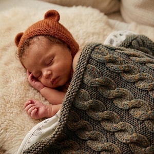 May include: A sleeping baby wearing a brown knit bear hat, nestled under a cable-knit blanket. The blanket is a mix of green, brown, and blue tones. The baby is lying on a soft, cream-colored surface.