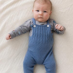 Puede incluir: Un bebé con un peto de punto azul claro con botones blancos y una camiseta de manga larga gris con un estampado. El bebé está acostado sobre una manta blanca.