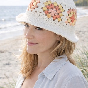 Puede incluir: Un sombrero de cubo de ganchillo blanco con un diseño floral en tonos melocotón, amarillo y rosa. El sombrero tiene un ala ancha, ideal para los días soleados. Un accesorio de moda.