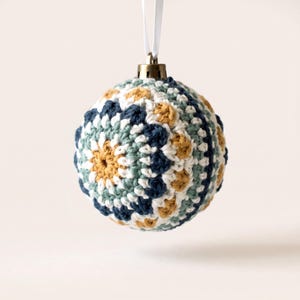 May include: A spherical Christmas ornament covered in a crocheted mandala pattern featuring concentric circles of mustard yellow, sage green, navy blue, and white yarn. The ornament is topped with a metallic cap and a white ribbon loop for hanging.