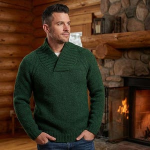 May include: A man wearing a dark green sweater with a shawl collar. The sweater is a solid color and appears to be made of a knit material. The background includes a fireplace with a lit fire and a wooden cabin interior.