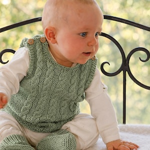 Puede incluir: Un bebé con un chaleco de punto verde salvia con diseño de punto de cable y botones de madera. El bebé también lleva patucos de punto a juego y una camisa y pantalón blancos de manga larga. El bebé está sentado sobre una superficie blanca.