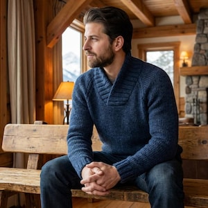 May include: A man wearing a blue sweater and jeans sits on a wooden bench near a window overlooking a snowy mountain range. A fireplace with a burning fire is in the background.