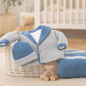 Puede incluir: Un conjunto de ropa de bebé de punto en una cesta de mimbre. El conjunto incluye una rebeca gris y azul, un gorro azul y gris y una manta a rayas azules y blancas. También hay un mordedor de oso de madera.
