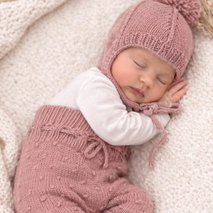 Puede incluir: Un bebé dormido con un gorro rosa tejido con pompón y pantalones a juego. El bebé también lleva una camiseta blanca de manga larga. El conjunto está hecho de hilo suave y texturizado, ideal para recién nacidos.