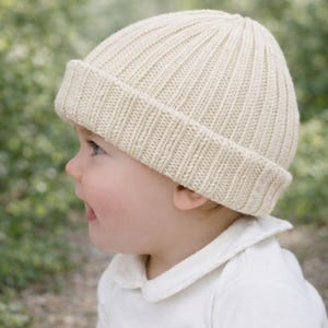 Puede incluir: Un gorro de punto acanalado de color crema con un borde doblado. El gorro está hecho de un hilo suave y texturizado y está diseñado para bebés o niños pequeños. El gorro se muestra en un bebé.