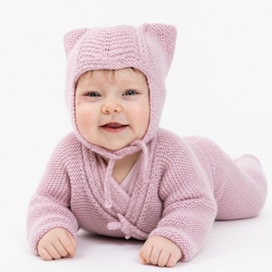 Puede incluir: Un conjunto de punto para bebé en color rosa empolvado que consta de una parte superior de manga larga estilo cruzado y pantalones a juego. El conjunto incluye un gorro a juego con dos pequeñas orejas puntiagudas y lazos finos de punto.