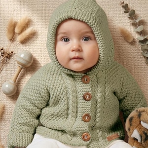 Puede incluir: Un bebé con una rebeca con capucha de punto verde salvia con botones de madera. La rebeca tiene un diseño de punto trenzado y una pequeña etiqueta que dice "COZY KNITS". El bebé está tumbado sobre una superficie beige.