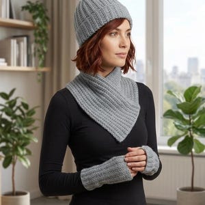 Puede incluir: Un conjunto gris de gorro, cuello y guantes sin dedos de punto. Los accesorios de invierno de punto acanalado se muestran en una persona que lleva una camisa negra de manga larga. El gorro es estilo gorro.