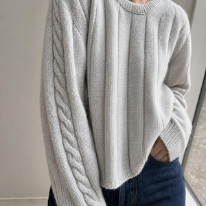 Puede incluir: Un suéter gris claro con la parte delantera acanalada y detalles de punto trenzado en las mangas. El suéter tiene cuello redondo y se combina con vaqueros azul oscuro. El estilo general es informal y cómodo.