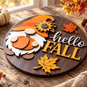 Puede incluir: Un letrero circular de madera con acabado de veta marrón oscuro que presenta un gnomo en capas con un sombrero naranja y un girasol, sosteniendo una calabaza, y el texto hello FALL en blanco y amarillo, con hojas de arce naranjas y amarillas.