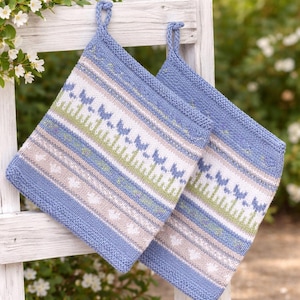 Op de afbeelding: Twee gebreide pannenlappen met een blauwe rand en ophanglus. Het ontwerp heeft strepen in wit, lichtgroen en beige, met blauwe vlinder- en hartmotieven. De pannenlappen leunen tegen een witte houten constructie.