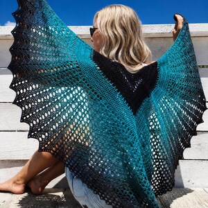Puede incluir: Un chal triangular de ganchillo con un degradado de color que va del negro sólido en el centro a un turquesa vibrante y vuelve al negro en los bordes festoneados. El intrincado patrón calado crea una textura delicada y encajada.