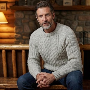 Puede incluir: Un suéter de punto trenzado gris claro con cuello redondo. El suéter tiene un patrón texturizado y se usa con jeans azules. La persona está sentada en un banco de madera en un ambiente rústico y acogedor.