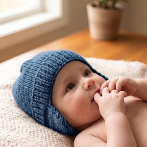 Puede incluir: Un bebé con un gorro azul tejido con orejeras. El bebé está acostado sobre una manta color crema, con los dedos en la boca. El gorro es de color liso y parece hecho a mano.