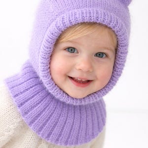 Puede incluir: Un gorro de punto lila con orejas de gato. El gorro está diseñado para cubrir la cabeza y el cuello, con un cuello acanalado. El niño lleva un suéter blanco. El gorro es un accesorio acogedor para el clima frío.