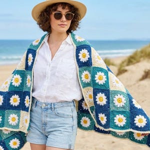Puede incluir: Un chal de ganchillo colorido con un diseño de margaritas en tonos azules, turquesa, crema y amarillo. El chal está drapeado sobre los hombros. El fondo muestra una escena de playa con arena y el océano.