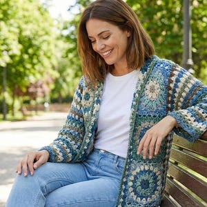Puede incluir: Una mujer con un cárdigan de ganchillo hecho a mano y abierto por delante. El cárdigan presenta un diseño de patchwork con tonos de azul, verde y beige. Lleva una camiseta blanca y vaqueros azules, sentada en un banco del parque.