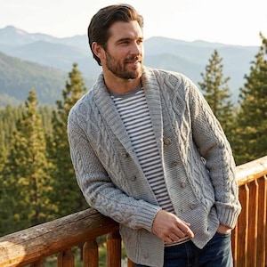 Puede incluir: Un cárdigan de punto trenzado gris claro con cuello chal y cierre de botones. El cárdigan se lleva sobre una camisa a rayas. El fondo presenta una barandilla de madera y una vista panorámica de un bosque y montañas.