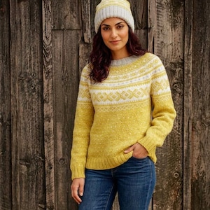 Puede incluir: Una mujer con un suéter amarillo y gorro a juego. El suéter tiene un patrón geométrico blanco en el pecho y las mangas. El gorro es de color amarillo claro y gris. Lleva vaqueros azules.