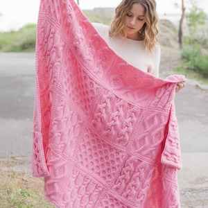 May include: A large, pink knitted blanket with a variety of textured patterns, including cable knit and dotted designs. The blanket is held up, showcasing its size and intricate details. The blanket appears to be made of soft yarn.
